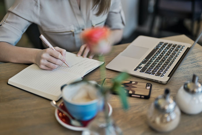 a woman writing something into her notebook lying near the laptop