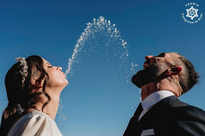 Couple in wedding attire laughing as water arcs between them under a clear sky.