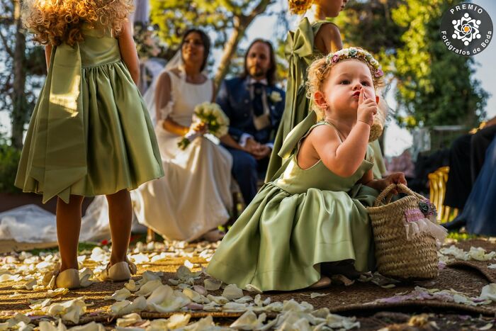 Child in green dress at wedding, making a shushing gesture, with couple and petals in the background.