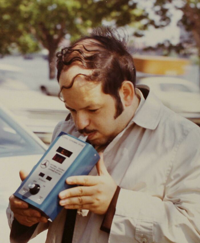 Man with unusual hair experimenting with a hand-held device outdoors.