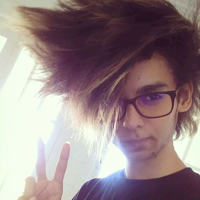 Young individual with a dramatic hair accident poses with glasses and a peace sign, showcasing unique hairstyle.