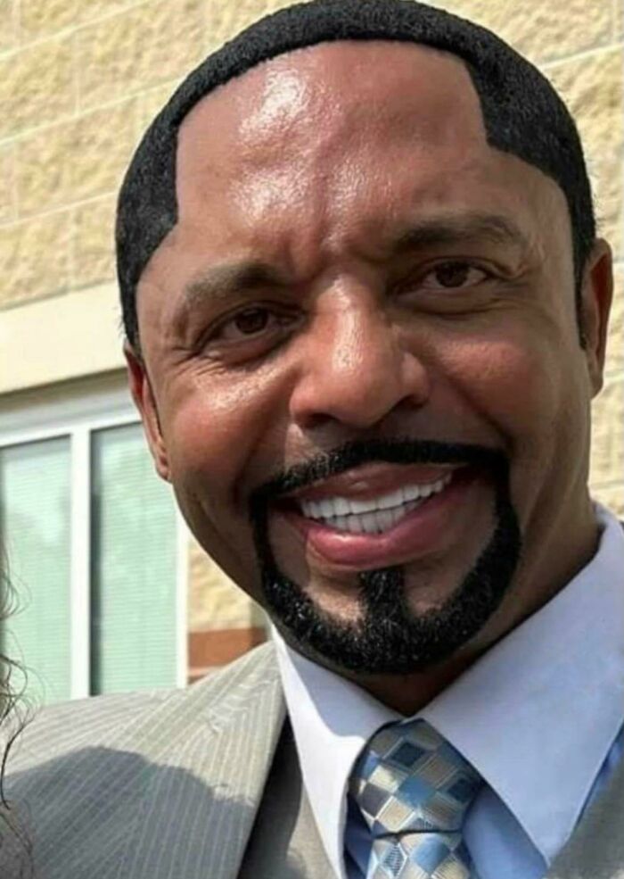 Man with a visibly unusual hairstyle and neatly trimmed beard, smiling in a suit and tie outdoors.