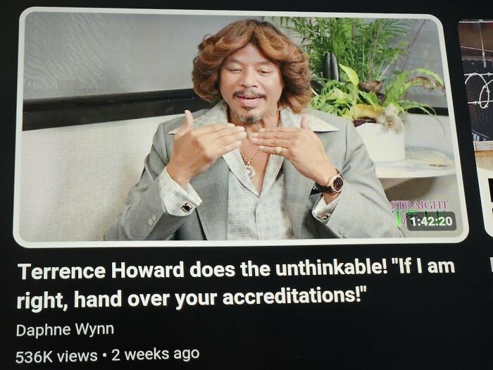 Person discusses hair accident, gesturing while wearing a gray suit in a room with plants and decor.