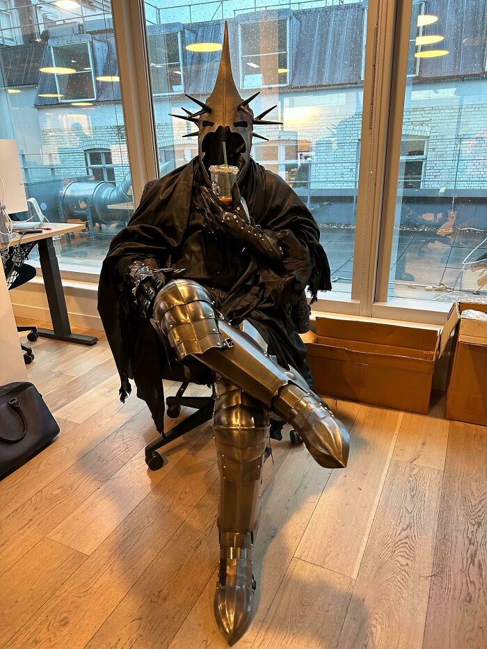 A person in a medieval knight costume holding a drink, sitting casually in an office with large windows.