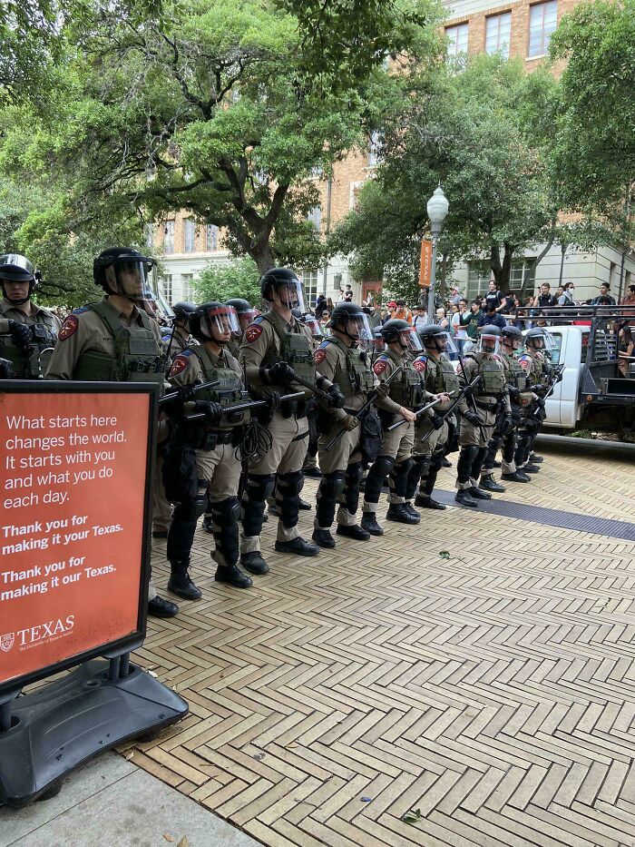 A line of armored officers stands in formation near a university sign, suggesting a boring dystopia.