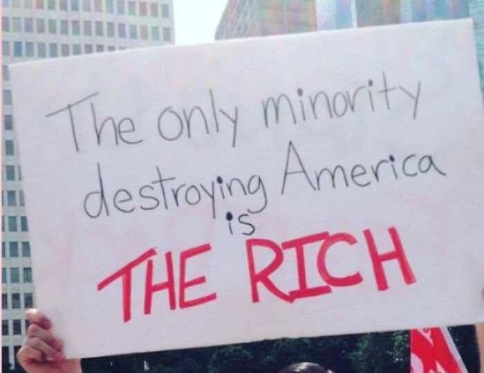 Protest sign reading "The only minority destroying America is the rich" held up against a cityscape background.