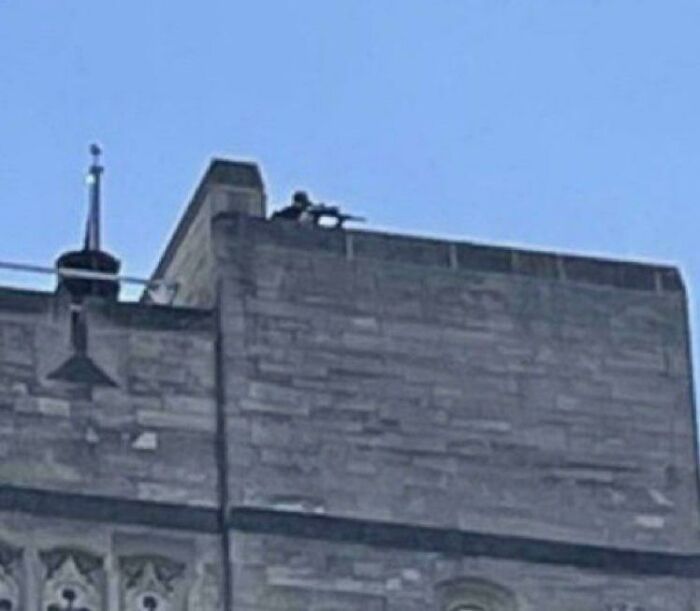 Rooftop observer in a dystopian setting, equipped with surveillance tools on a stone building against a clear blue sky.