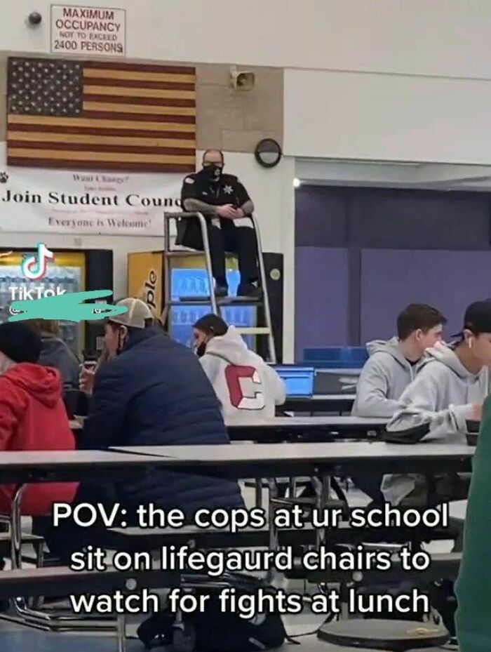 School cafeteria scene with a cop on a lifeguard chair watching students, showcasing peak dumb humor.