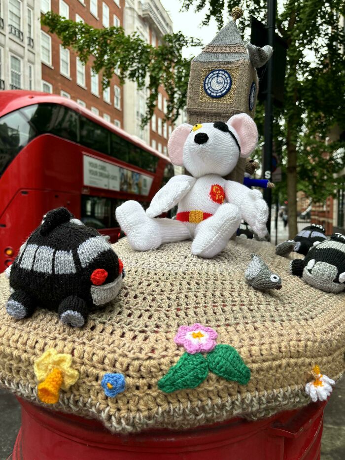 Knitted mouse with Big Ben hat on a mailbox, with red bus in the background, capturing a British scene.