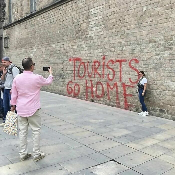 People taking photos in front of "Tourists Go Home" graffiti on a city wall, illustrating humans of capitalism pictures.