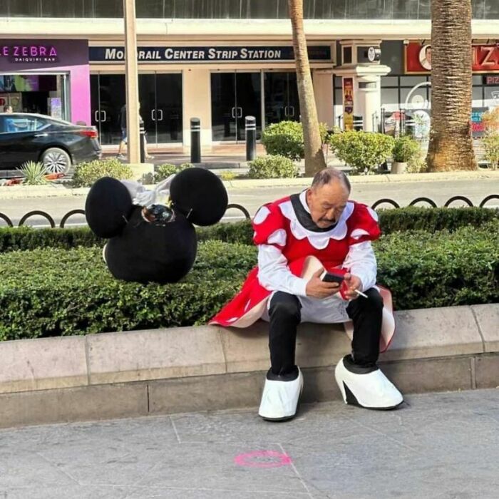 Man in a costume sitting on a bench, checking his phone, beside a large character head on a sunny sidewalk.