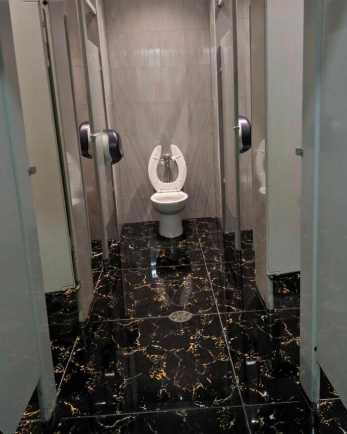 Marble bathroom with poor design—a toilet in a narrow stall with glossy, dark tile flooring. Design disaster example.