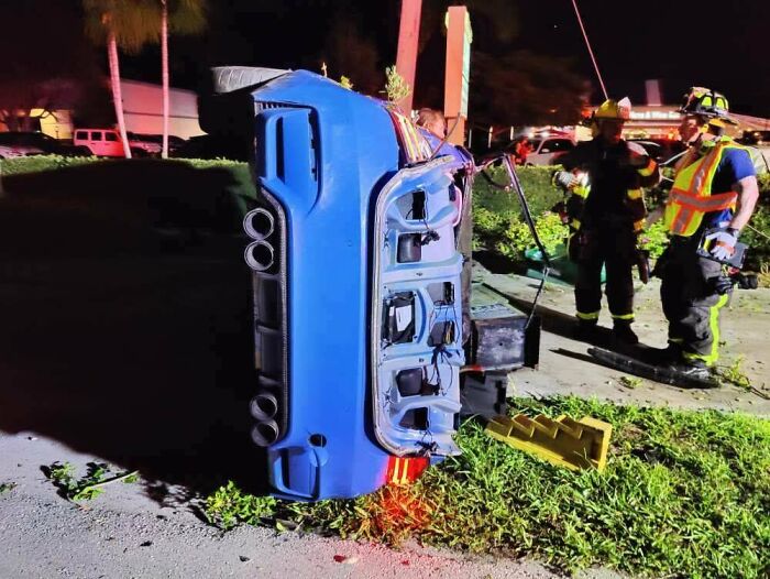 Overturned blue car on the side of the road with emergency responders nearby; an unfortunate incident.