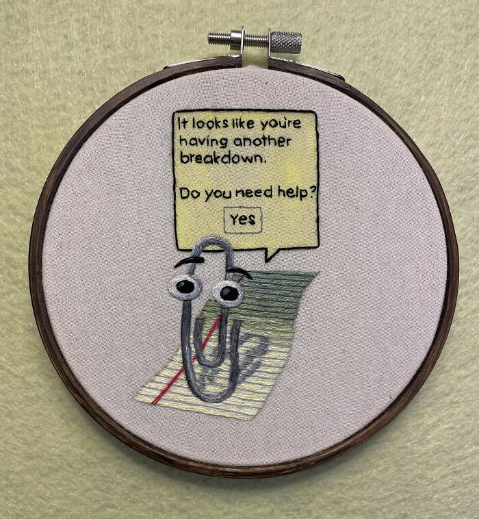 Creative embroidery featuring Clippy on a notepad with humorous text asking for help, displayed in an embroidery hoop.