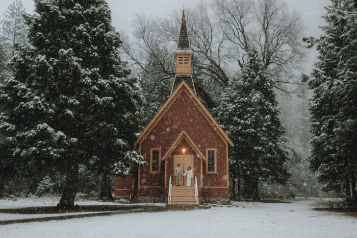 A couple poses in front of a rustic chapel in a snowy forest, showcasing top destination wedding photography 2024.