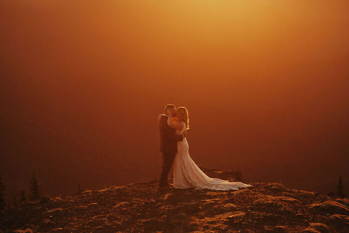 Couple embracing during a destination wedding at sunset, on a mountain, with a warm orange sky backdrop.