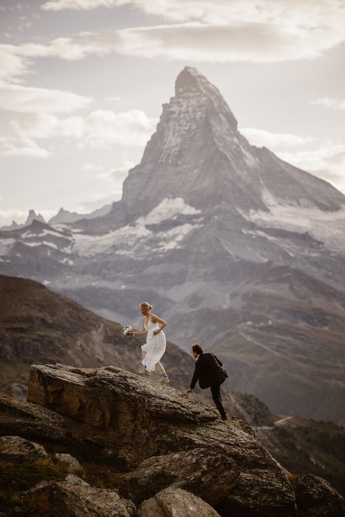 Couple in wedding attire climbing rocks with mountain backdrop, capturing a stunning destination wedding photo.