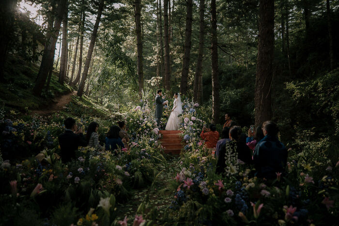 Couple exchanging vows in a forest setting, surrounded by guests and vibrant flowers.