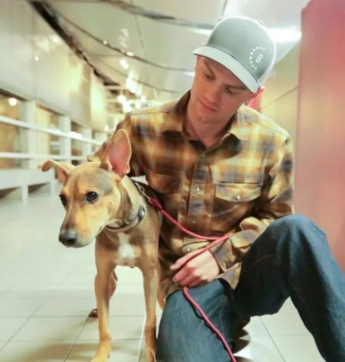 Man with a rescued dog on a red leash, wearing a plaid shirt and cap, sitting indoors.