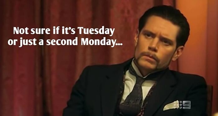 A man in a suit sitting at a table with a sign that says "not sure if it's Tuesday or just a second Monday".