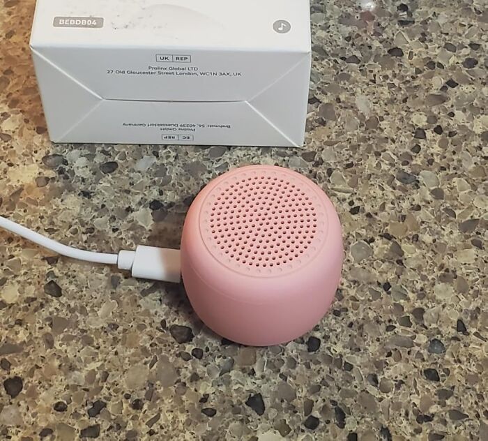 Pink mini speaker on granite counter, ideal for minimalist carry-on packing, with charging cable and white box in background.