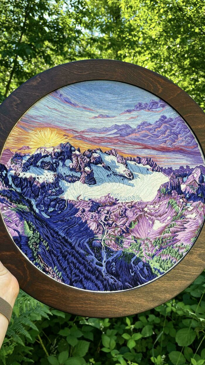 Creative embroidery depicting a colorful mountain landscape in a circular wooden frame, held outdoors.