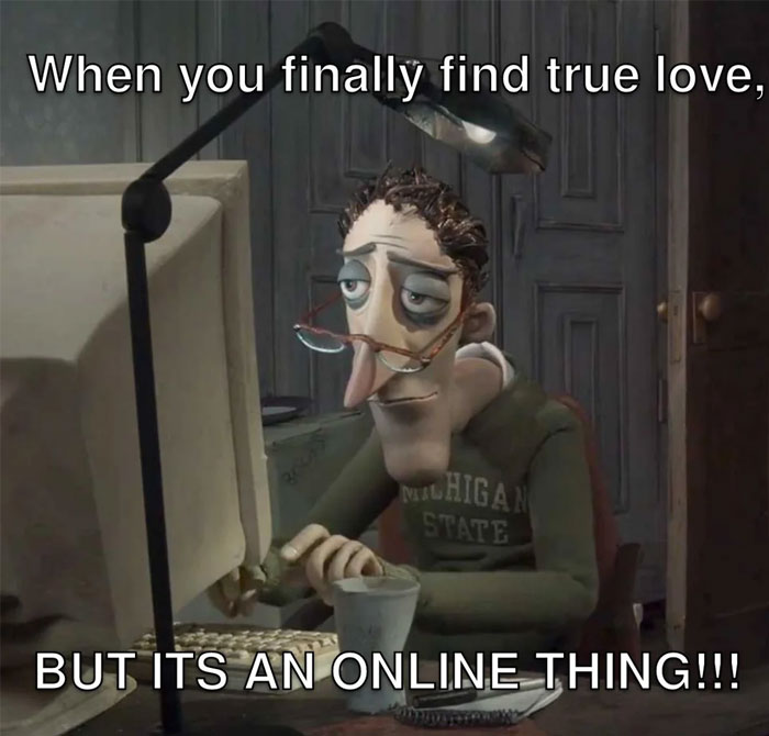 A love meme featuring a tired-looking animated character with glasses, wearing a Michigan State sweatshirt, sitting at a desk in front of a computer under a desk lamp. The text above the image reads, "When you finally find true love," and below it says, "BUT IT'S AN ONLINE THING!!!"