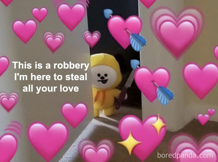 A love meme featuring a plush toy character dressed in a yellow costume, peeking from behind a wall. The background is filled with pink heart emojis and blue arrows. The text on the image reads, "This is a robbery, I'm here to steal all your love." The setting appears to be indoors with carpeted flooring.