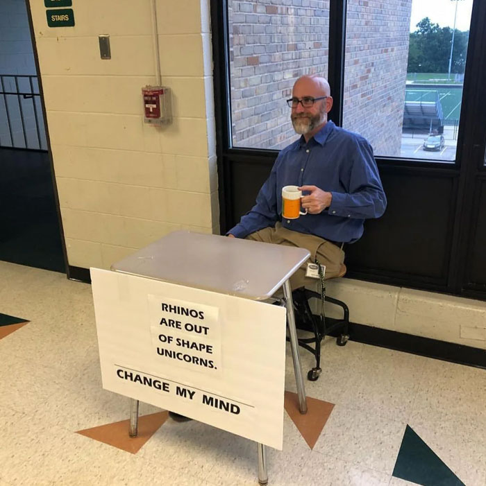 Meme of the day: A man sits at a small desk in a hallway with a sign taped to the front. The sign reads, "Rhinos are out of shape unicorns. Change my mind." The man is holding a mug and smiling, creating a humorous scene that invites debate.