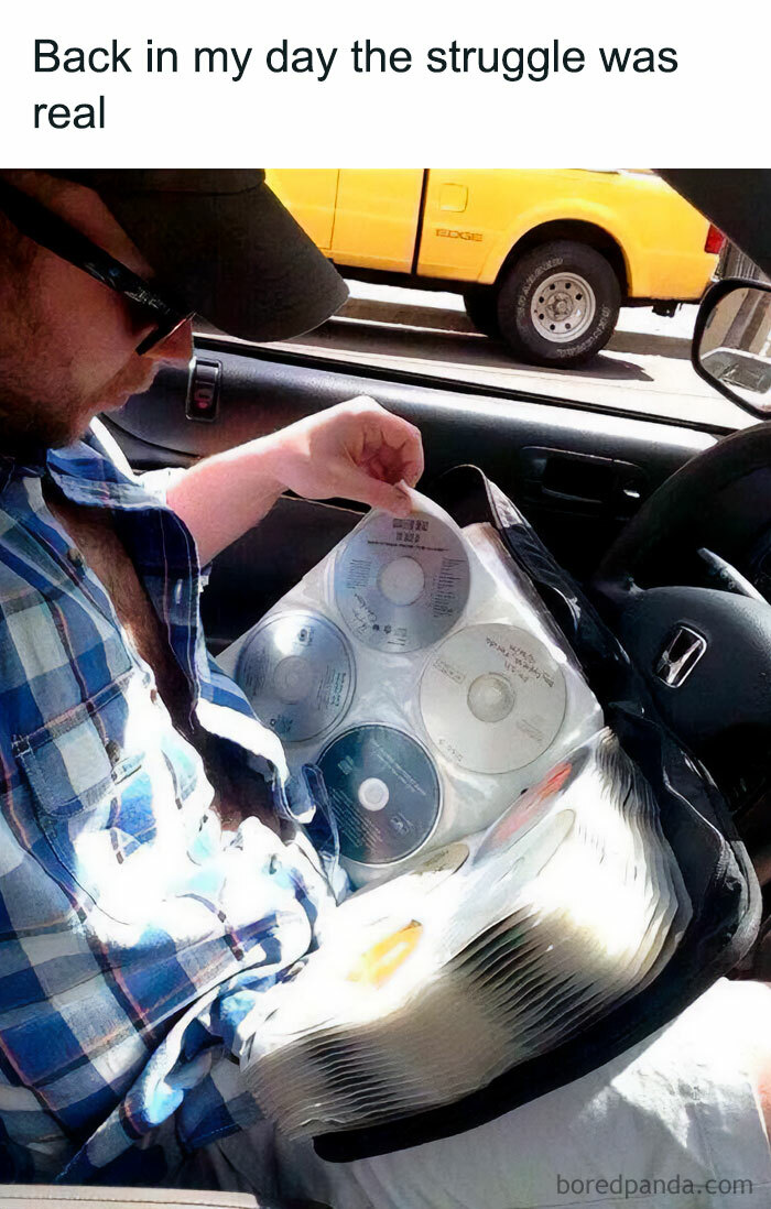 Meme of the day: The image shows a man sitting in a car, flipping through a large CD binder filled with discs. He is wearing a baseball cap and a plaid shirt, focusing on selecting a CD. The text above the image reads, "Back in my day the struggle was real," humorously referencing the pre-digital era when people had to manually choose and switch CDs for music in their cars.