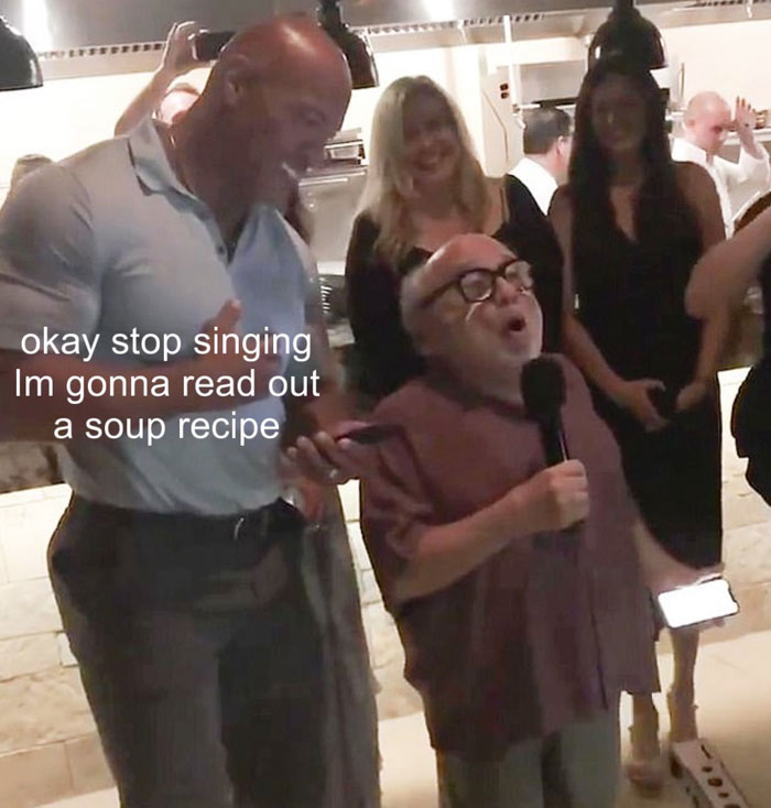 A The Rock meme featuring Dwayne "The Rock" Johnson standing next to Danny DeVito at a party. The Rock is smiling while DeVito holds a microphone, appearing to be passionately speaking or singing. The text on the image reads, "okay stop singing I'm gonna read out a soup recipe," humorously suggesting that instead of performing, DeVito is about to recite a soup recipe, adding an absurd and comedic twist to the scene.