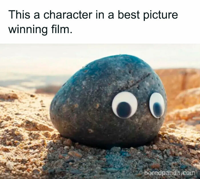 A The Rock meme showing an image of a small rock with googly eyes placed on a sandy surface, resembling a playful character. The text above reads, "This a character in a best picture winning film," humorously referencing the simplicity of the rock compared to high-profile movie characters, with a subtle nod to The Rock's nickname and persona.