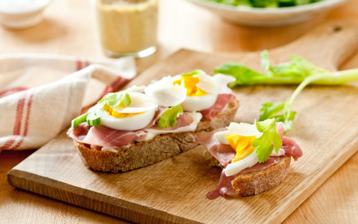 A Lunch idea featuring two slices of rustic bread topped with ham, soft-boiled egg halves, and garnished with celery leaves, served on a wooden board.