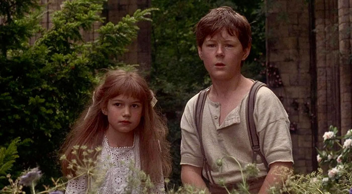 A young boy and girl from “The Secret Garden” movie stand outdoors in a lush garden, looking ahead with curious and slightly concerned expressions. The girl wears a white lace dress, and the boy is dressed in a simple shirt with suspenders. They are surrounded by greenery and flowers, with an old stone structure visible in the background. The scene conveys a sense of mystery and adventure, reminiscent of a period setting.