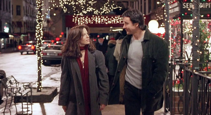 A scene from the movie Serendipity featuring a man and a woman walking side by side on a festive city street at night. Both are dressed in winter coats, with the woman wearing a red scarf, as they exchange smiles and conversation. The street is adorned with holiday lights, and storefronts, including a decorated café, add to the warm, romantic ambiance. In the background, cars and pedestrians contribute to the lively city atmosphere.