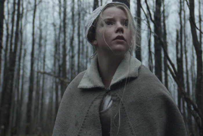 A scene from the movie The Witch featuring a young woman in period clothing, including a bonnet and a gray woolen cloak, standing in a gloomy, leafless forest. She gazes upward with a tense, fearful expression as if sensing danger around her. The barren trees and muted tones of the setting create an eerie, unsettling atmosphere.