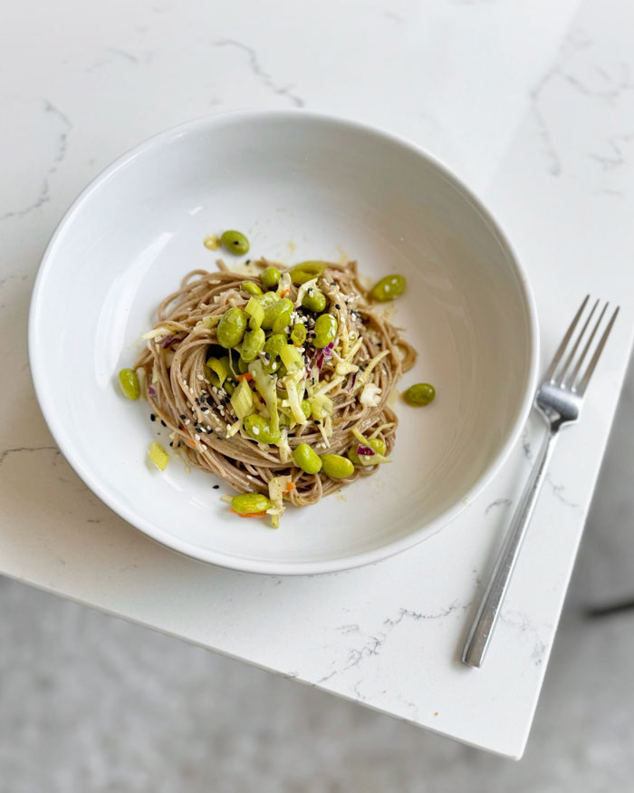 A lunch idea featuring sesame soba noodles topped with edamame, cabbage, and sesame seeds, served in a white bowl on a marble table.