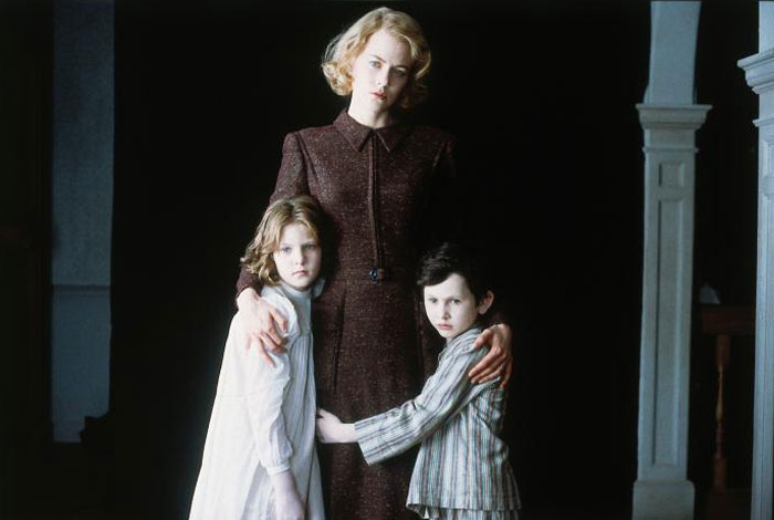 A scene from the movie The Others featuring a woman in a dark, vintage dress standing protectively with her arms around two young children, a girl and a boy. The children cling closely to her, both wearing nightclothes, and have solemn, anxious expressions. The background is dark, creating a haunting and mysterious atmosphere, with the woman looking serious and watchful.