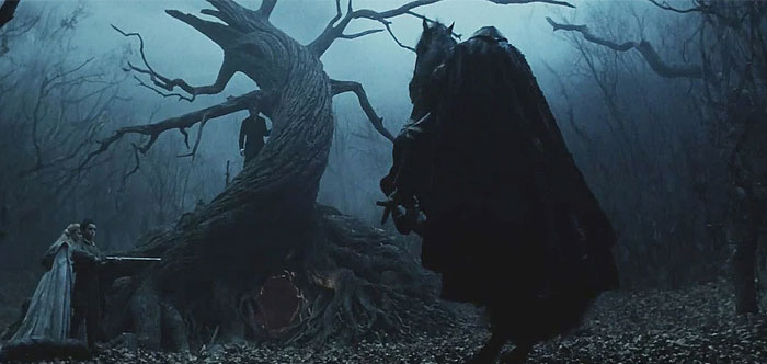  A dark, eerie scene from the movie Sleepy Hollow featuring a cloaked figure on horseback, presumably the Headless Horseman, standing before a twisted, gnarled tree in a fog-filled, leafless forest. A large, ominous opening is visible at the base of the tree, and in the background, two figures can be seen cautiously approaching, adding to the suspenseful atmosphere. The scene is shrouded in deep shadows, enhancing its supernatural and foreboding tone.