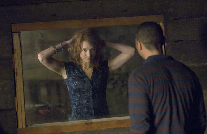 A scene from the movie The Cabin in the Woods featuring a young woman adjusting her hair in front of a mirror, while a man stands on the other side, watching her through what appears to be a one-way window. The setting is inside a rustic, wooden cabin, creating a tense and mysterious atmosphere. The woman seems unaware of being observed, while the man looks intently, heightening the feeling of secrecy and unease.