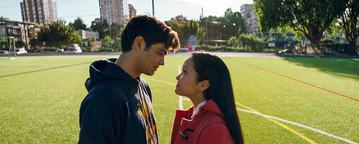 A scene from the movie To All the Boys I've Loved Before featuring a young man and woman standing face-to-face on a bright, sunlit sports field. The man, wearing a hoodie, gazes intently at the woman, who is dressed in a red coat. Both have serious yet affectionate expressions as they share a quiet, intimate moment. The backdrop includes trees, buildings, and the lines of the field, adding to the peaceful, outdoor setting.