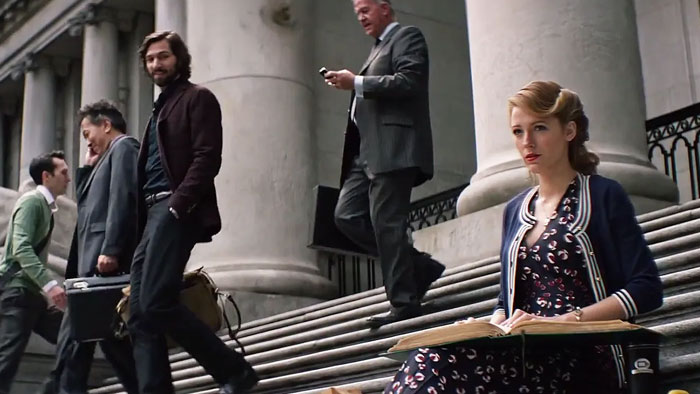 A scene from the movie The Age of Adaline featuring a young woman sitting on the steps of a grand building, dressed in a vintage-style outfit with a cardigan and patterned dress, holding an open book. She has a contemplative expression as she observes her surroundings. Several people, dressed in modern business attire, walk down the steps behind her, including a man checking his phone. The scene contrasts the character’s timeless style with the contemporary hustle around her.