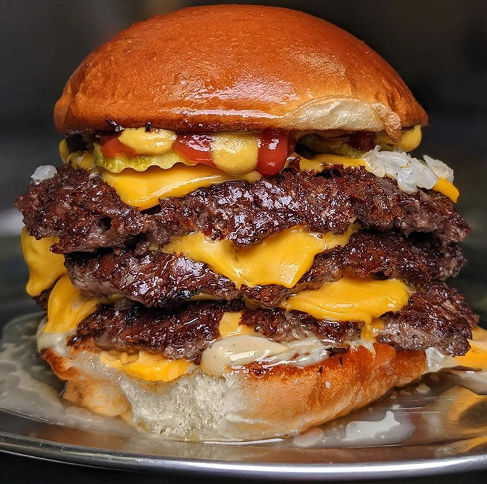 A Lunch idea featuring a stacked smash burger with multiple layers of crispy beef patties, melted cheese, pickles, onions, ketchup, and mustard on a soft bun.