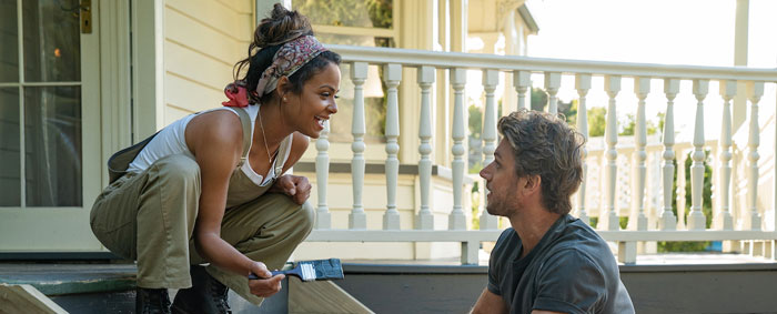 A scene from the movie Falling Inn Love featuring a woman and a man working together on a home renovation. The woman, crouching on the steps of a porch, holds a paintbrush and smiles warmly at the man. She is dressed casually in overalls and a headscarf. The man, sitting nearby, looks up at her with a smile, creating a lighthearted and cheerful atmosphere. The backdrop includes the porch of a cozy house, adding to the scene’s sense of charm and teamwork.