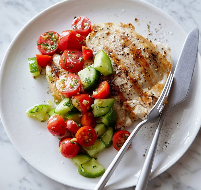 A Lunch idea featuring a grilled chicken breast served with a fresh cucumber and cherry tomato salad, garnished with dill on a white plate.