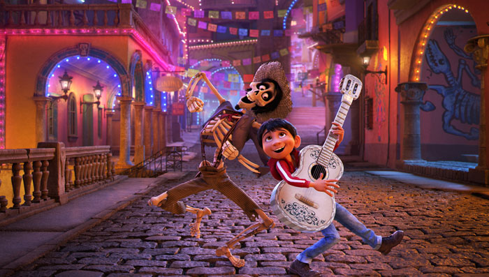 A lively scene from the movie Coco featuring a young boy holding a white guitar with intricate designs and a skeleton wearing a hat, both joyfully dancing on a cobblestone street. The background shows colorful buildings adorned with lights and papel picado banners, creating a festive atmosphere in the Land of the Dead.