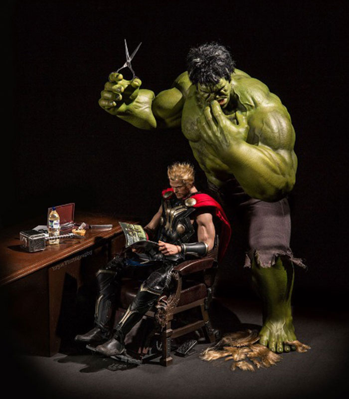 An action figure scene featuring Hulk standing behind Thor, who is sitting in a chair reading a magazine. Hulk is holding a pair of scissors above Thor’s head, looking focused as if about to give him a haircut. Thor appears relaxed and unaware of Hulk’s actions. 