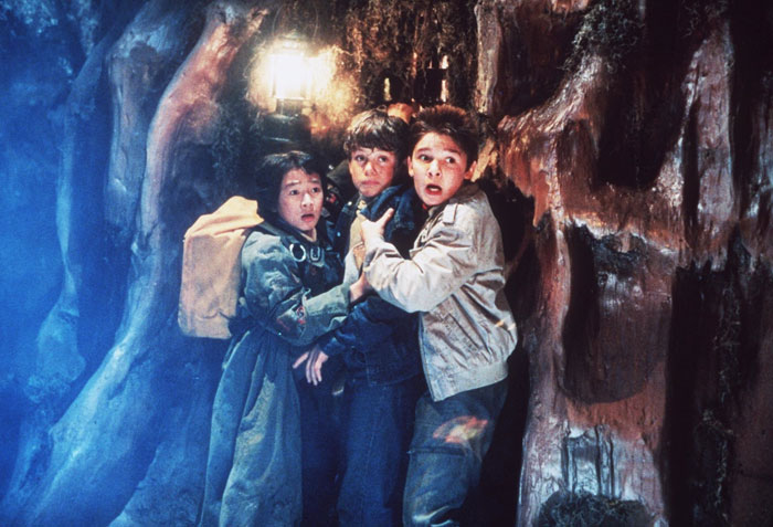 A tense scene from the movie The Goonies featuring three young boys huddled together in a narrow cave passage, looking frightened and alert. A lantern glows behind them, casting dim light on the rugged, damp walls. The boys wear jackets and backpacks, appearing lost and nervous as they brace for something unknown.