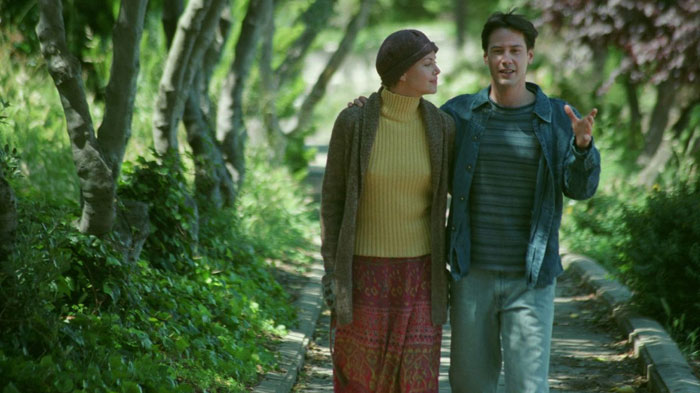 A scene from the movie Sweet November featuring a man and a woman walking arm-in-arm down a tree-lined path. The woman, wearing a headscarf, a yellow turtleneck, and a patterned skirt, looks affectionately at the man. The man, dressed casually in a jacket and jeans, gestures while talking, smiling as they walk together in a peaceful, green setting.