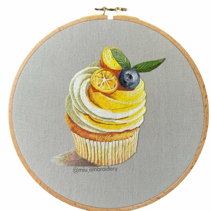 Creative embroidery of a cupcake topped with citrus slices and a blueberry, displayed in a wooden hoop.